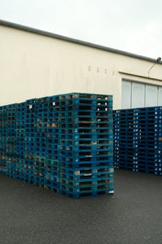 High stacks of blue plastic pallets outside a warehouse building.