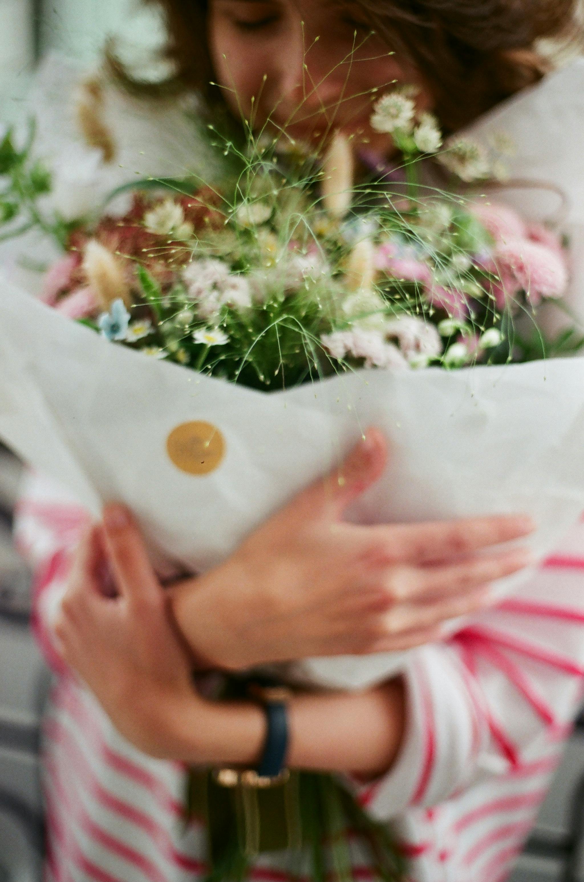 A woman in striped clothing embraces a vibrant bouquet of flowers with a soft, blurred background.
