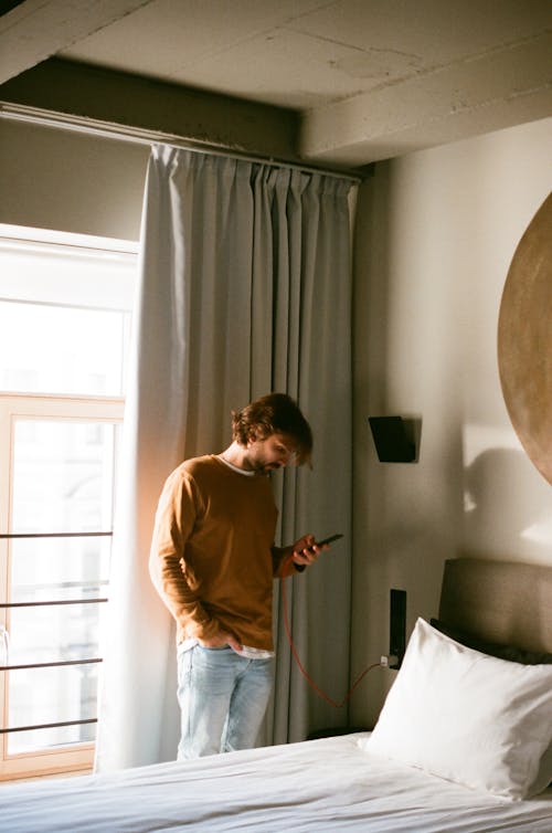 Man in Brown Sweater Using a Cellphone Beside a Bed