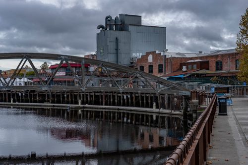Free Industrial bridge with reflection on river, bordered by historic buildings under a moody, cloudy sky. Stock Photo