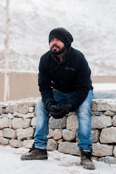 Bearded man in winter attire sitting on a stone wall during snow in India.