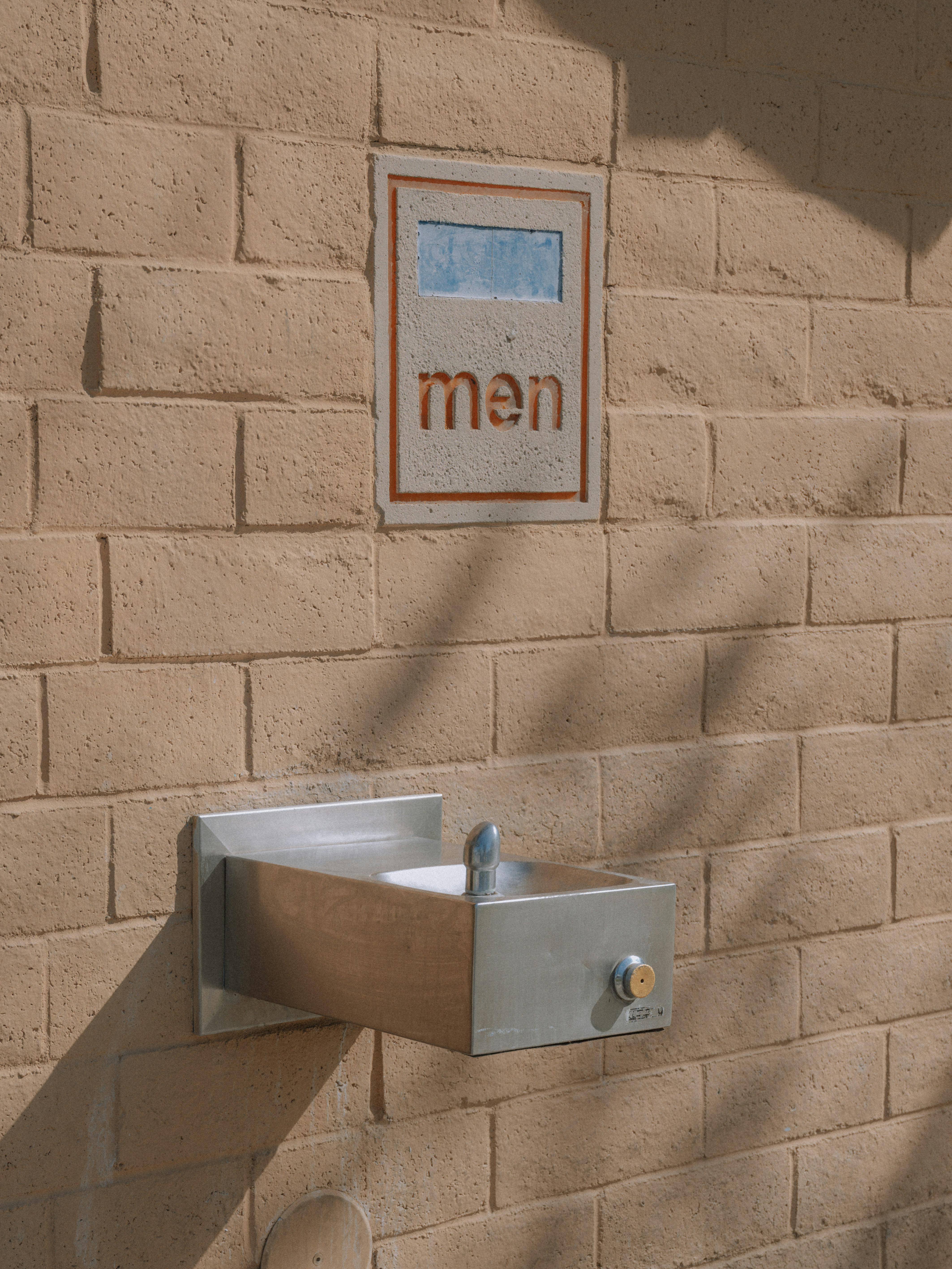 A public drinking fountain attached to a brick wall near a men's restroom sign.