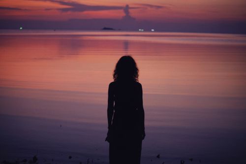Free A silhouette of a woman standing by a calm lake at sunset, creating a peaceful scene. Stock Photo