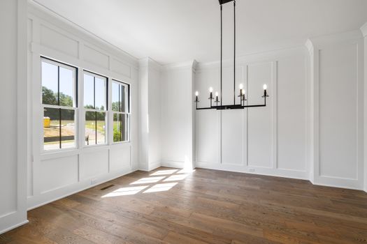 A spacious, light-filled room featuring white walls, wooden floors, and a modern chandelier.