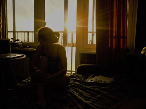A woman sits inside a sunlit room, reflecting by the window at sunset in St. Petersburg, Russia.