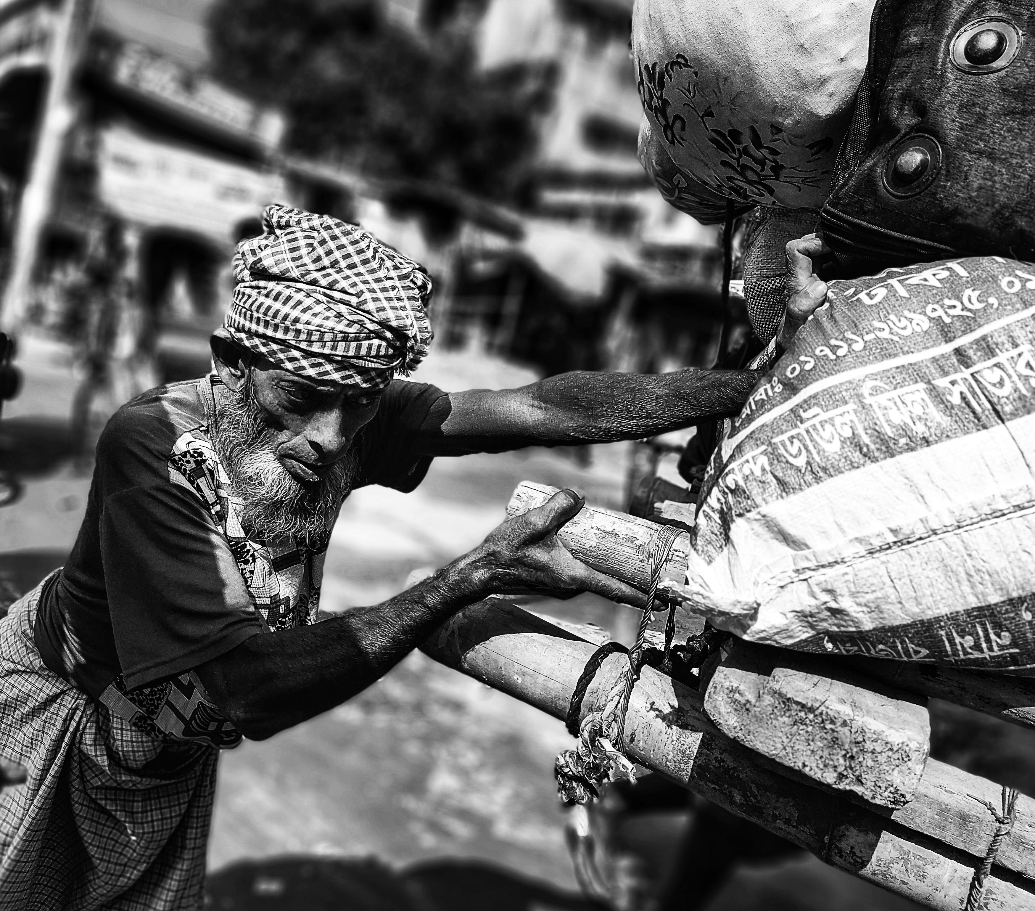 Elderly man pushing heavily loaded cart in Bangladesh street, captured in black and white.