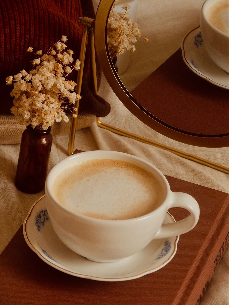 A warm coffee setup with a ceramic mug, dried flowers, and a mirror reflection.