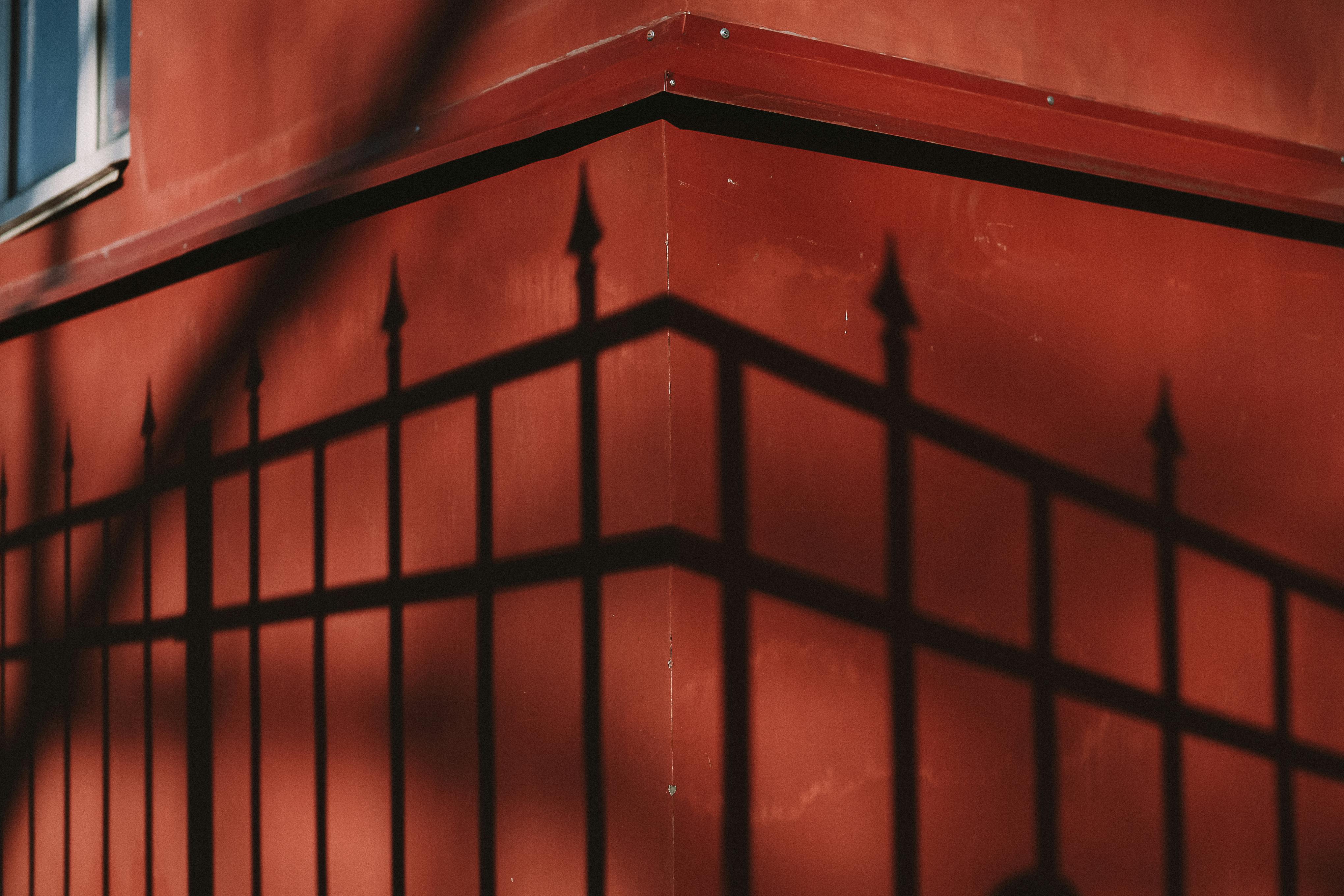Artistic shadow of iron fence cast on a vibrant red building corner, highlighting the interplay of light and architecture.