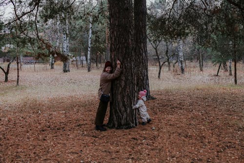 Woman in Brown Jacket and a Baby Hugging the Tree Trunk 