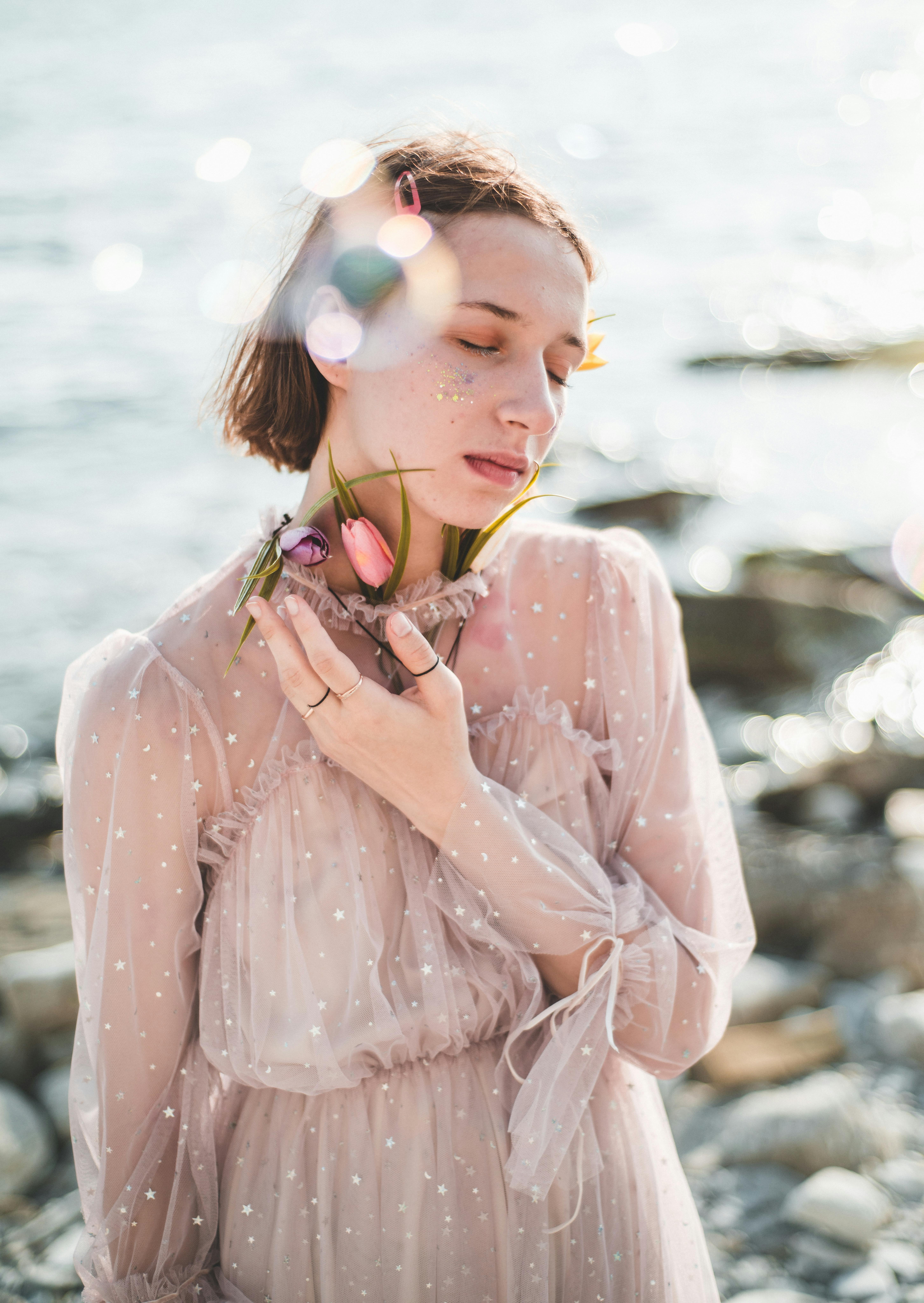 A serene woman with eyes closed, holding flowers, standing by the sunlit waterfront.