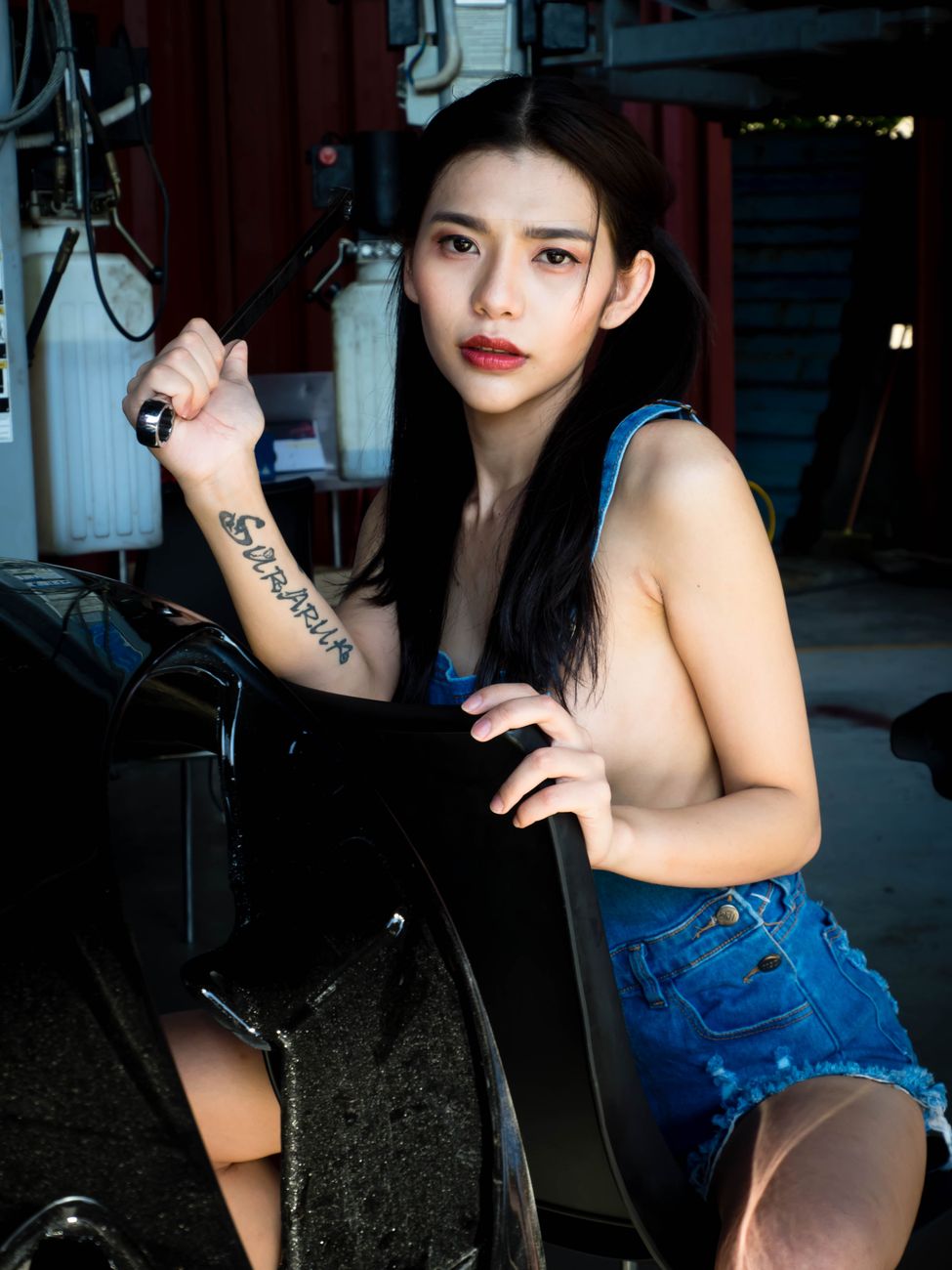 A tattooed woman in a denim jumper poses confidently with a tool in an auto workshop.