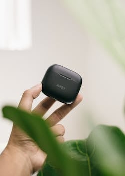 Close-up of a hand holding wireless earbuds case with greenery in the background.