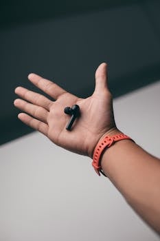 A hand with a red watch holds a black wireless earbud in a minimalist setting.