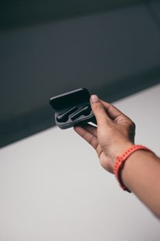 A hand with a red wristband holds a black wireless earbuds case against a grey background.