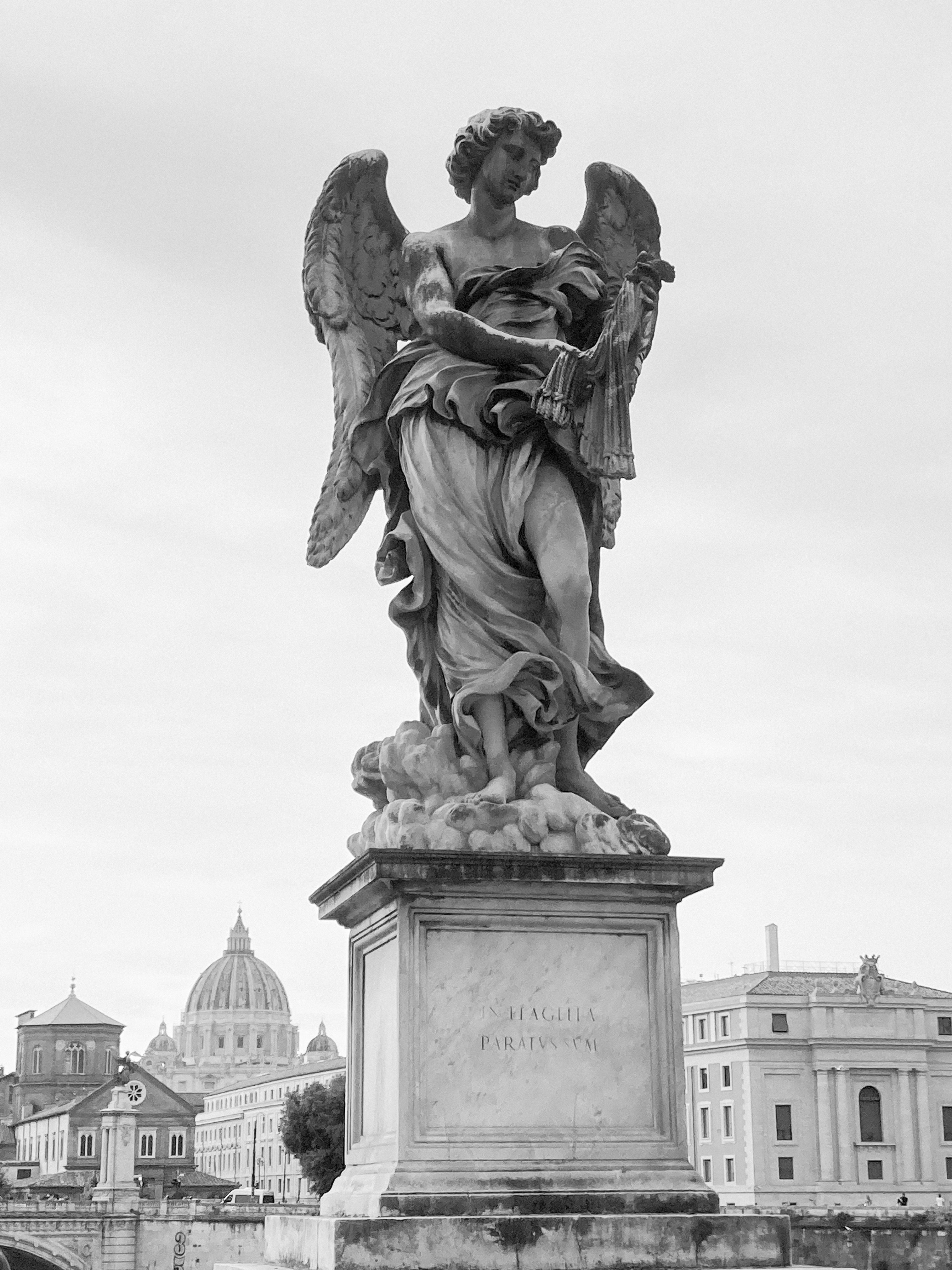 Grayscale image of an angel sculpture at Castel Sant'Angelo, Rome, with St. Peter's Basilica in the background.