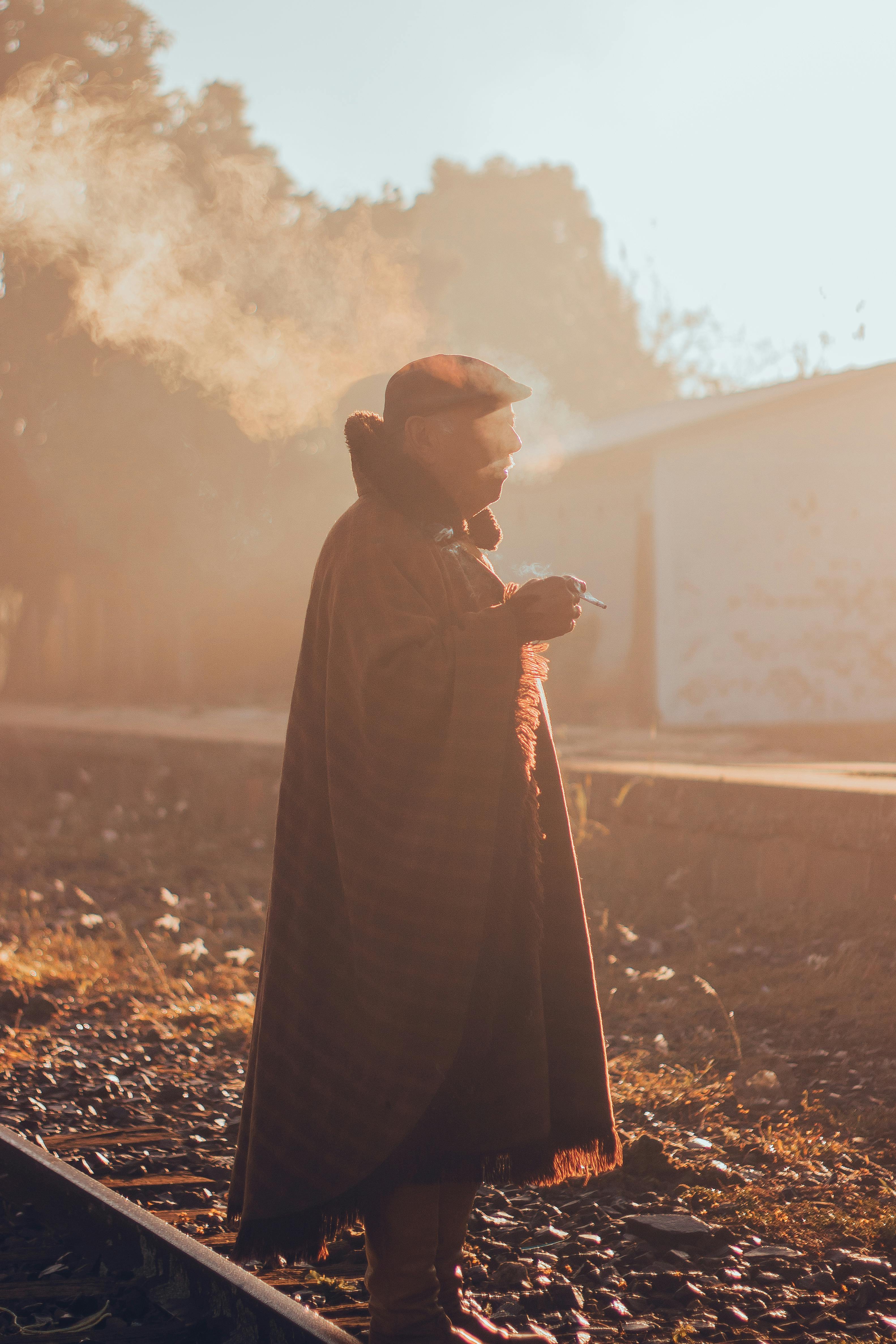 An elderly man in a cap smokes on a winter day by railway tracks with warm sunlight.