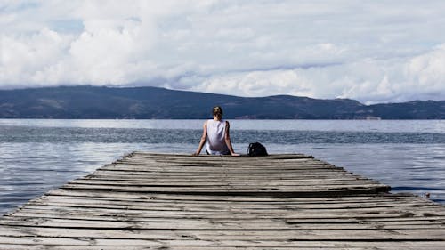 Person Sitting on Brown Wooden Dock Under Cloudy Blue Sky