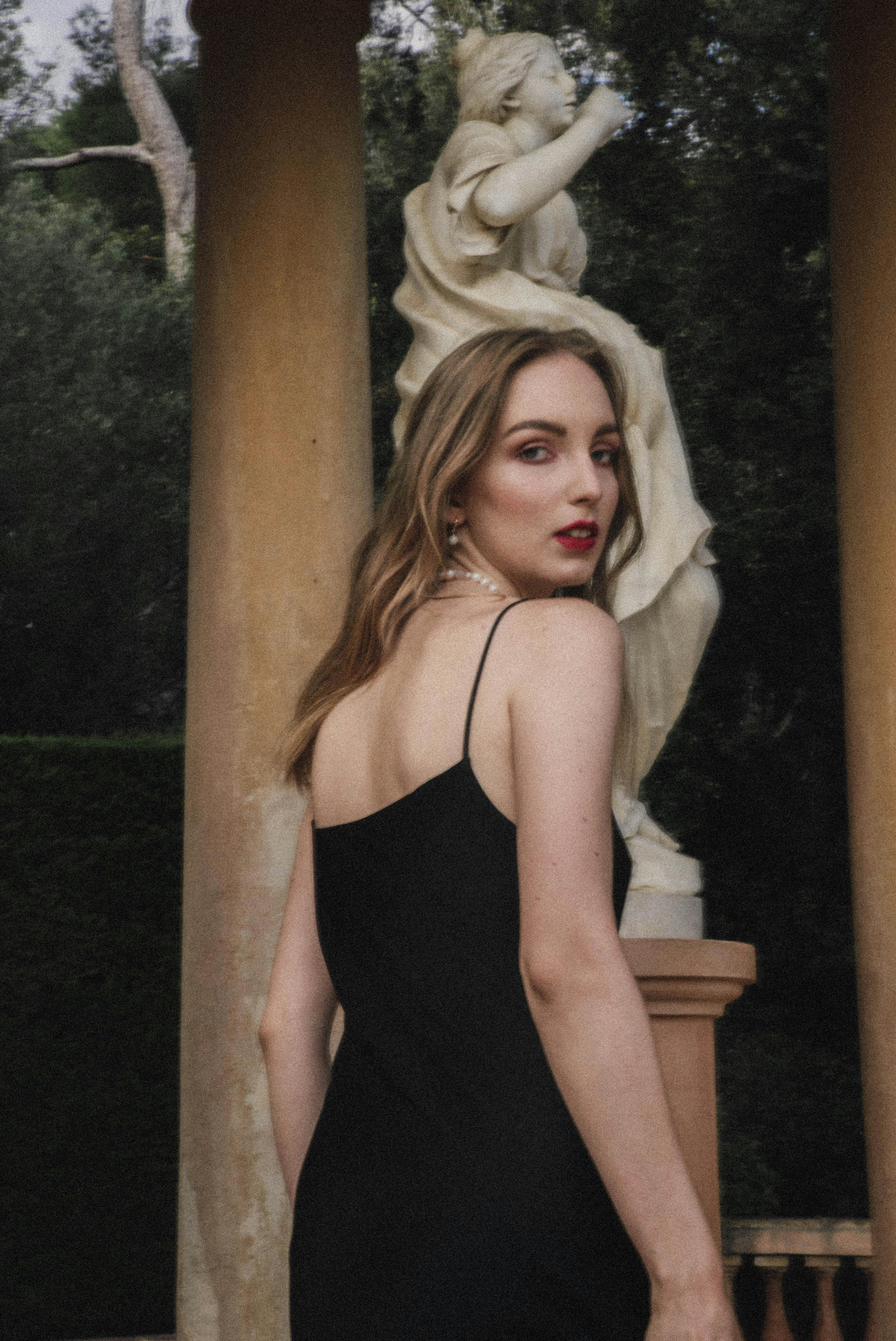 A woman in a black dress poses beside a classical statue outdoors.
