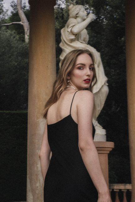 A woman in a black dress poses beside a classical statue outdoors.