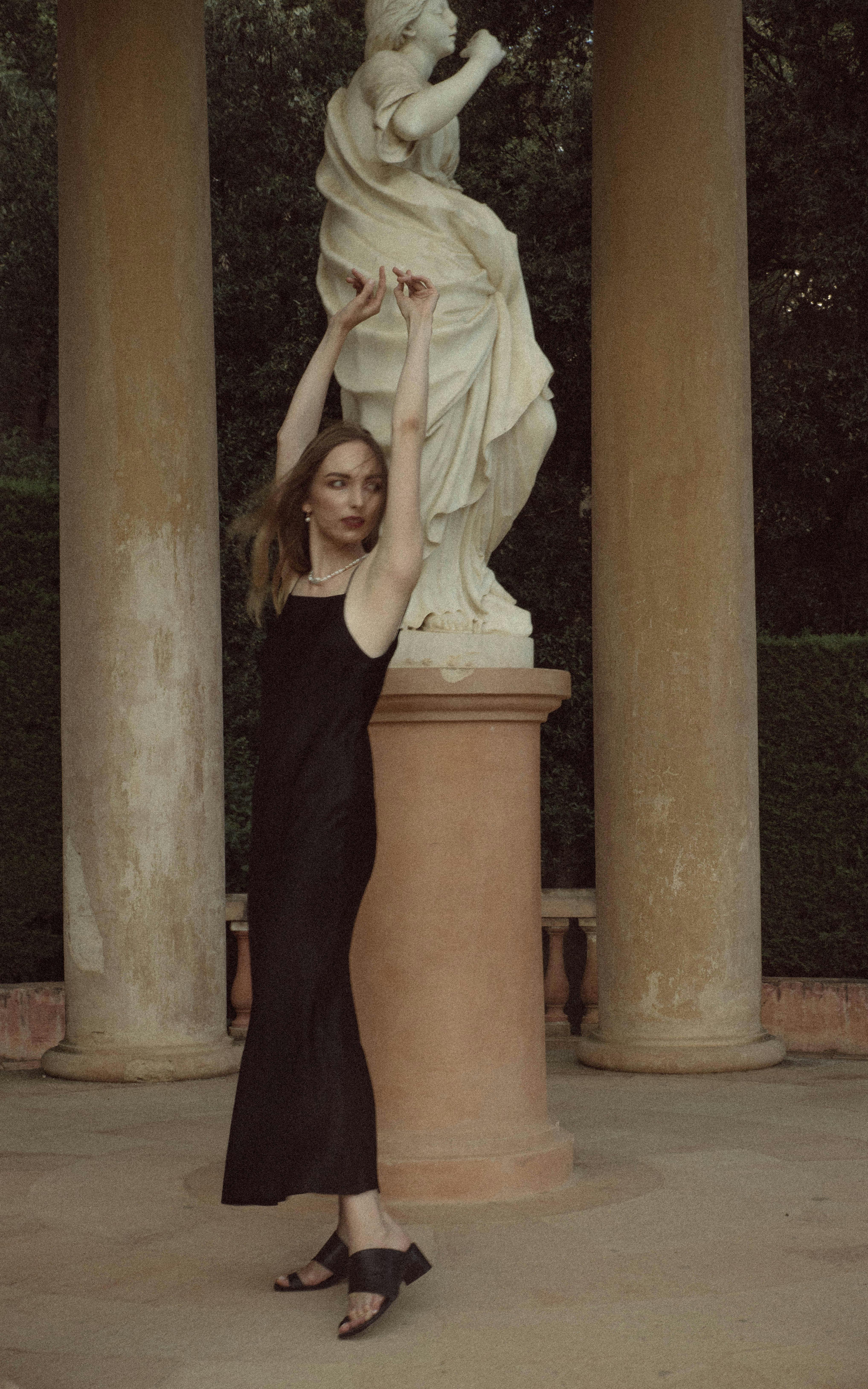 A woman in a sleek black dress poses elegantly next to a classical statue amidst columns.