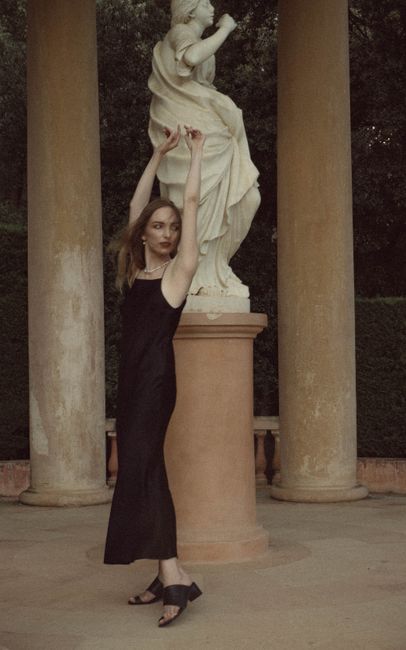 A woman in a sleek black dress poses elegantly next to a classical statue amidst columns.