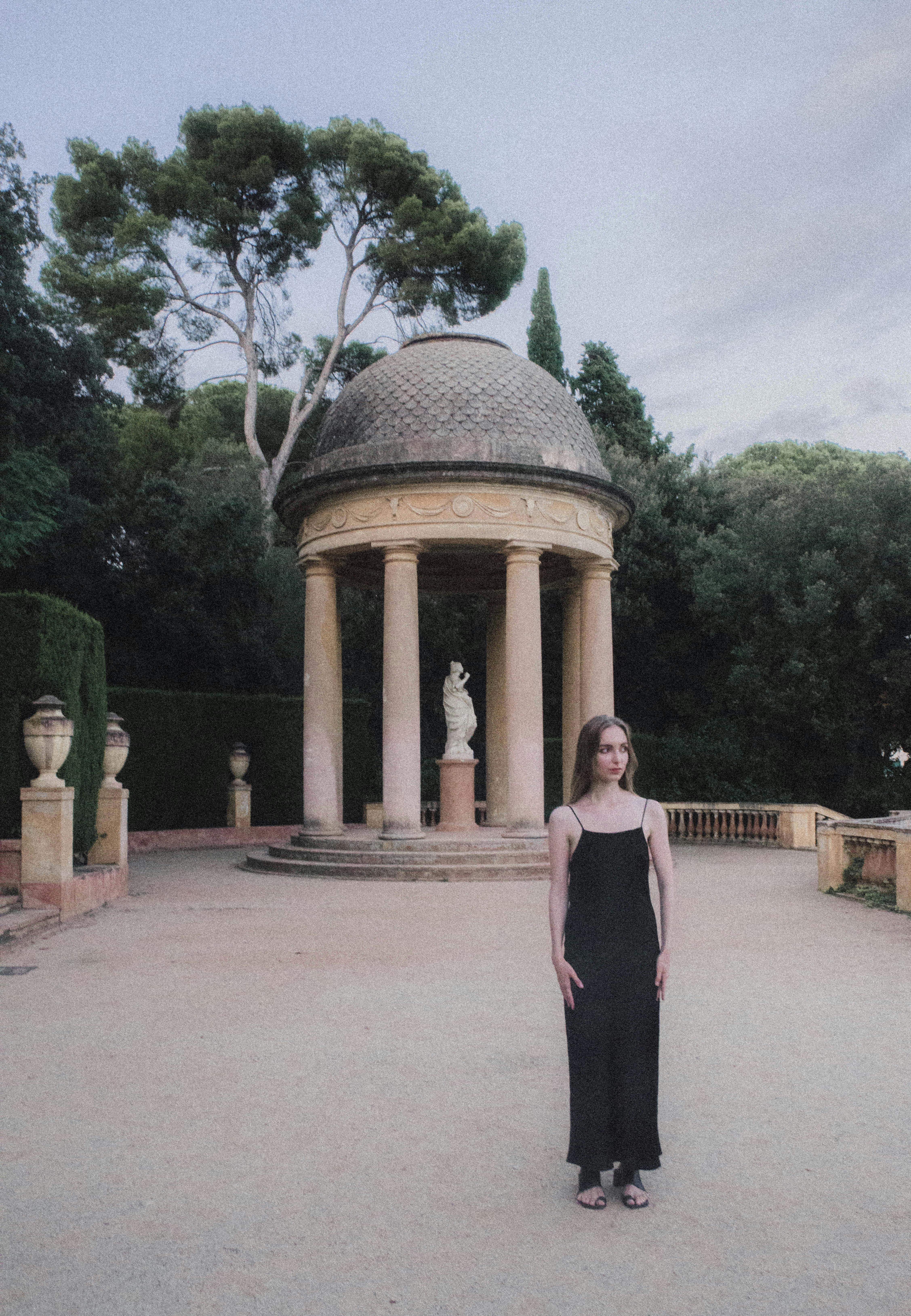 A woman in a black dress stands by a classical pavilion, blending elegance with timeless architecture.