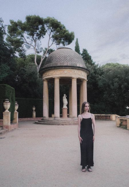 A woman in a black dress stands by a classical pavilion, blending elegance with timeless architecture.
