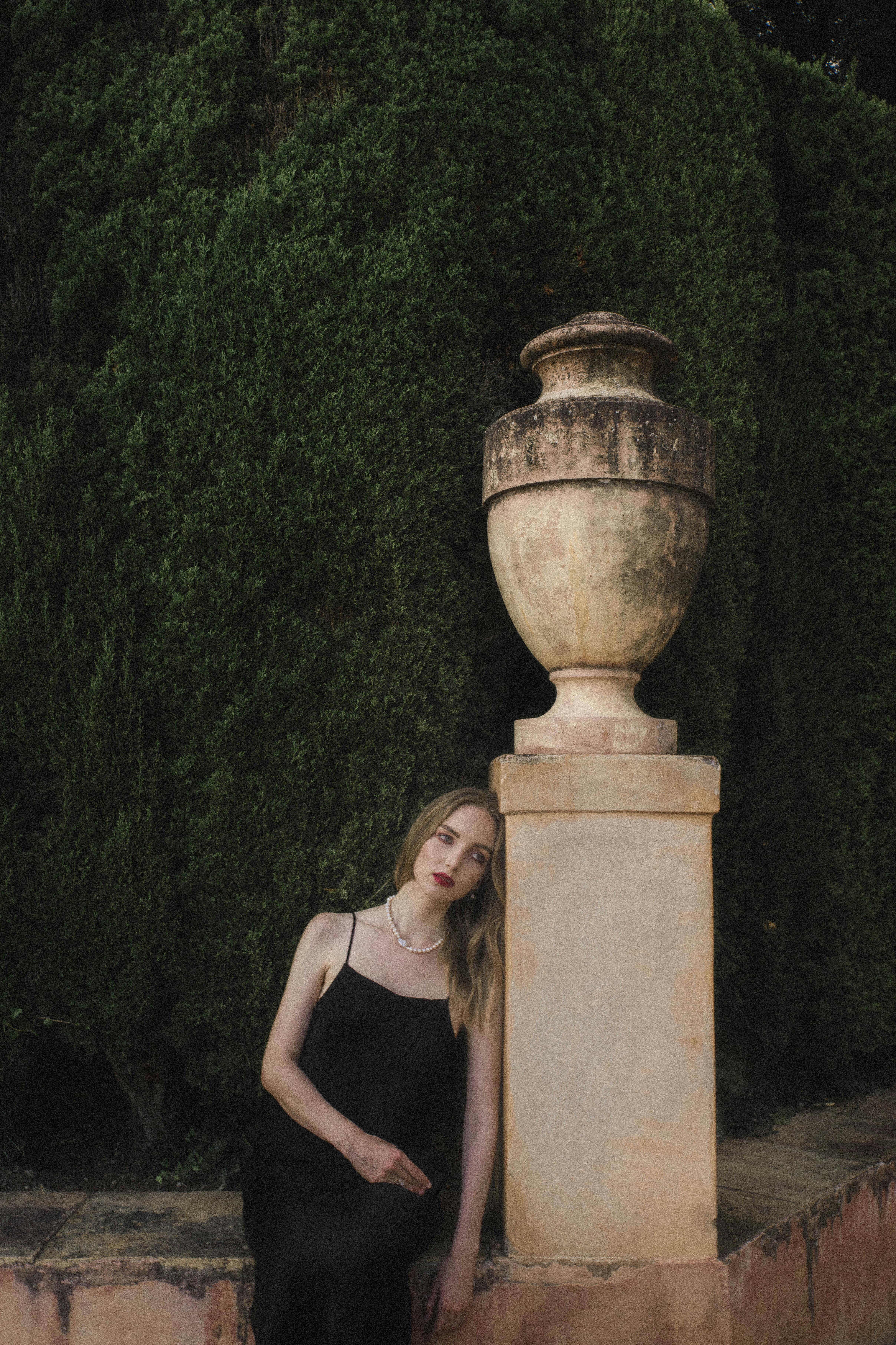 A woman in a black dress leaning against a vintage stone urn in a serene garden.