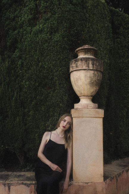 A woman in a black dress leaning against a vintage stone urn in a serene garden.