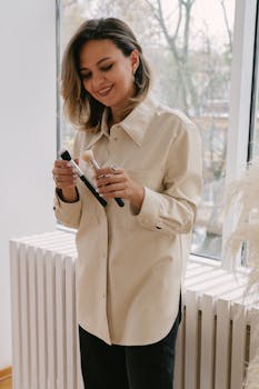 A smiling woman in a button-down shirt holds makeup brushes near a window, creating a serene indoor scene.