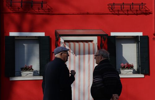 Free Two senior men talking in front of a bright red building with white accents and flower boxes. Stock Photo