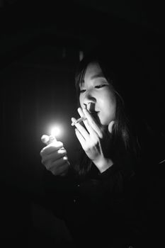 An East Asian woman lights a cigarette in a dramatic black and white setting.