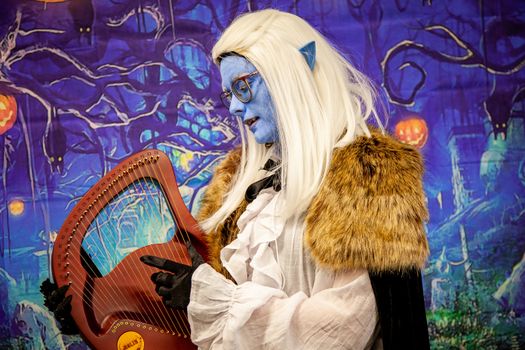 A fantasy-themed musician in elaborate costume playing a harp indoors against a mystical backdrop.