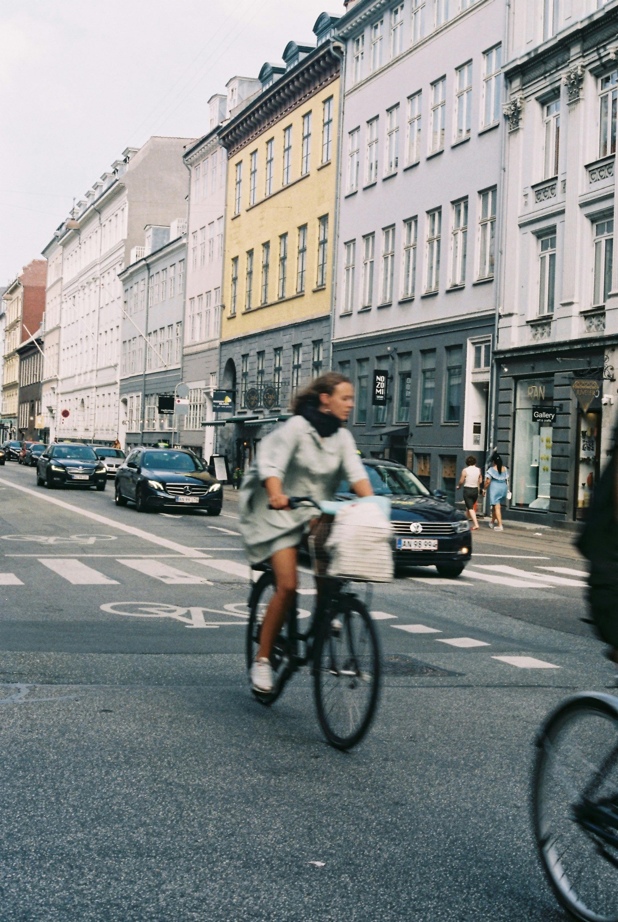 A woman rides a bicycle on a busy urban street, surrounded by cars and buildings.
