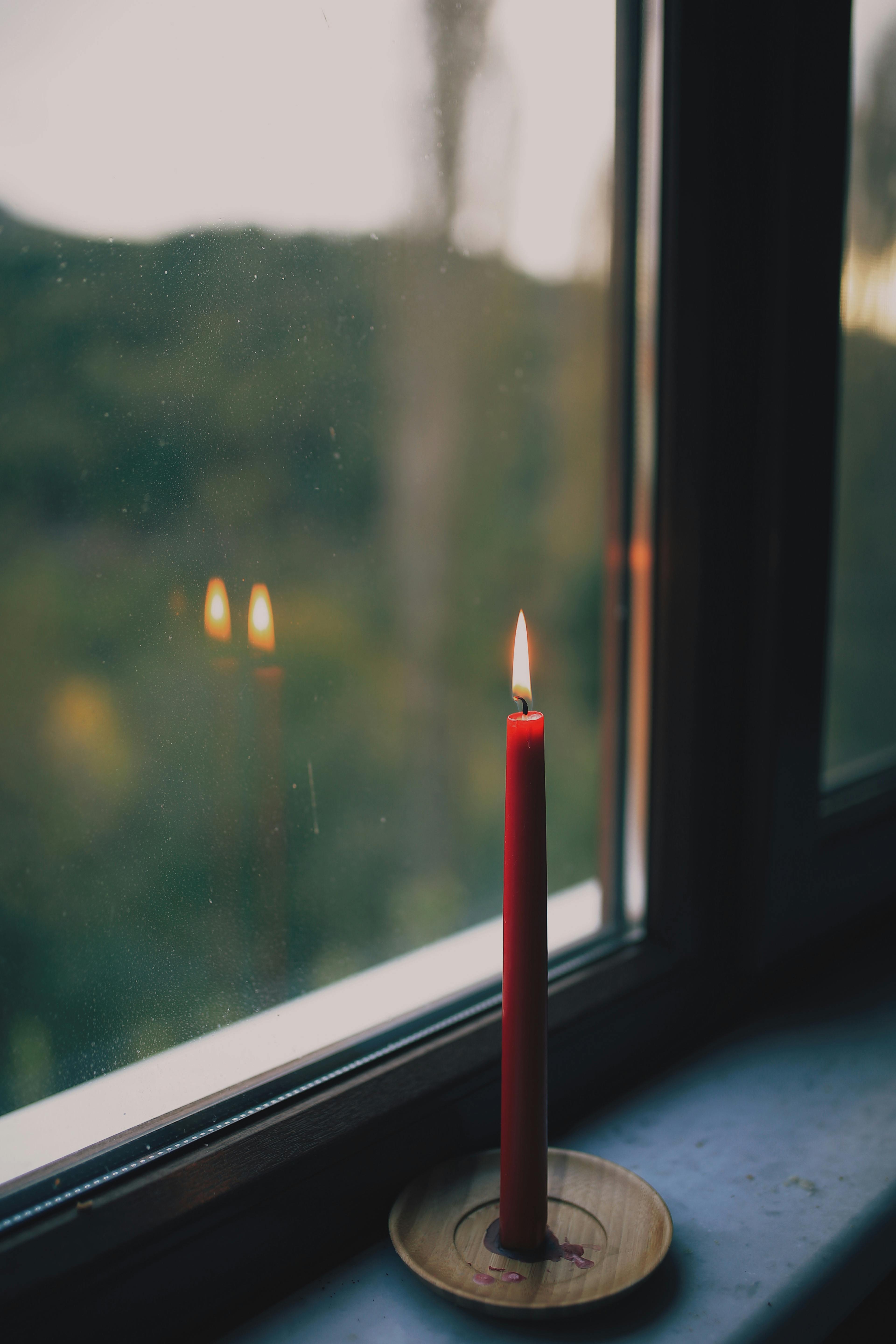 Close-up of a red candle burning on a windowsill with a forest view in the background.