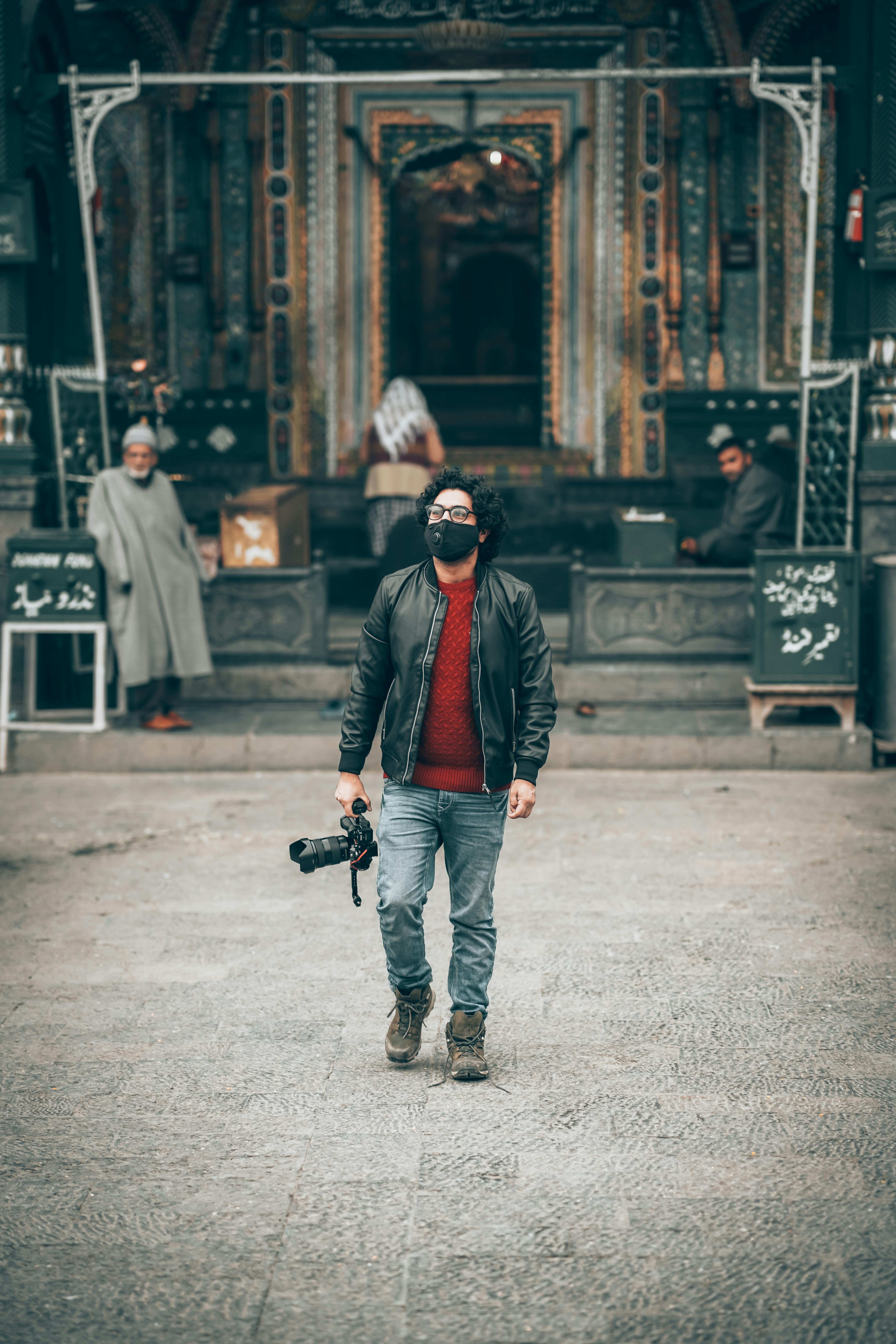 A photographer wearing a face mask and black jacket at a historic site holding a camera.