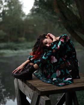 A woman in a floral dress poses thoughtfully on a wooden platform by the misty lake.