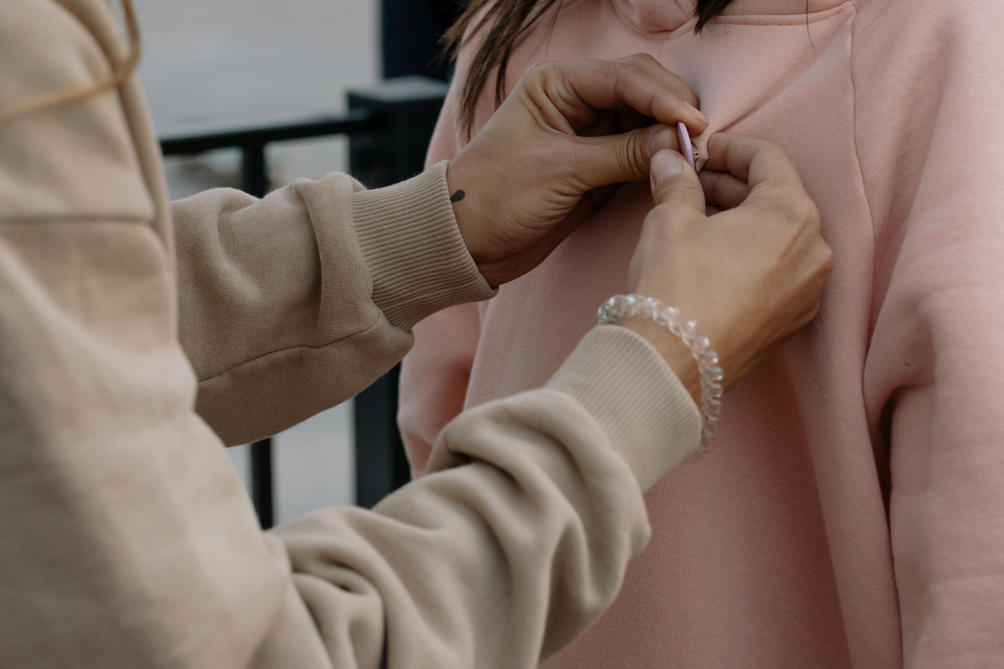 Close-up of hands pinning a badge on a pink sweatshirt outdoors.