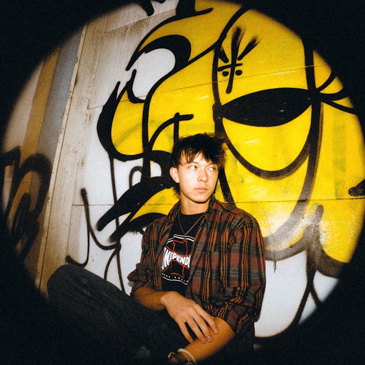 Young Man In Flannel Shirt Sitting By Wall With Graffiti