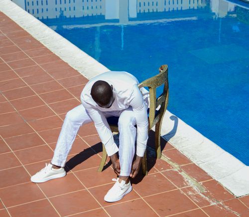 Free A man in white clothing sits on a chair by a swimming pool, adjusting his shoes on a sunny day. Stock Photo