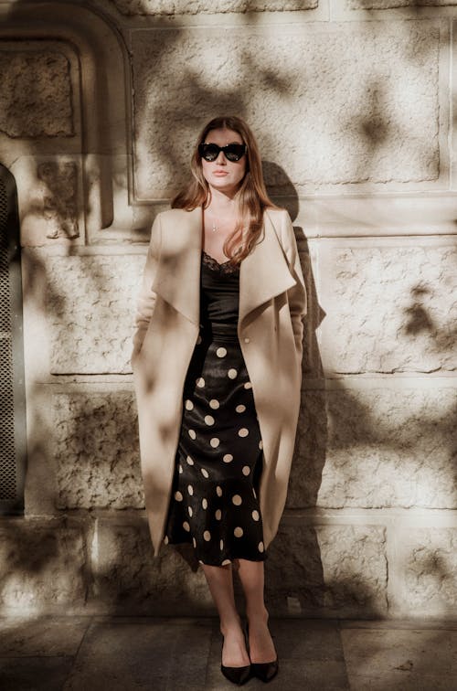 A Woman in Black Dress and Beige Coat Leaning on the Wall