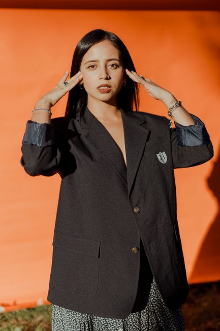 Young woman in stylish black blazer poses with bright orange background outdoors.
