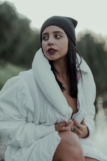 Young woman in a white jacket and knit hat sitting outdoors with a thoughtful expression.
