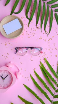 Stylish eyeglasses on a pink surface with fern leaves, alarm clock, and card.