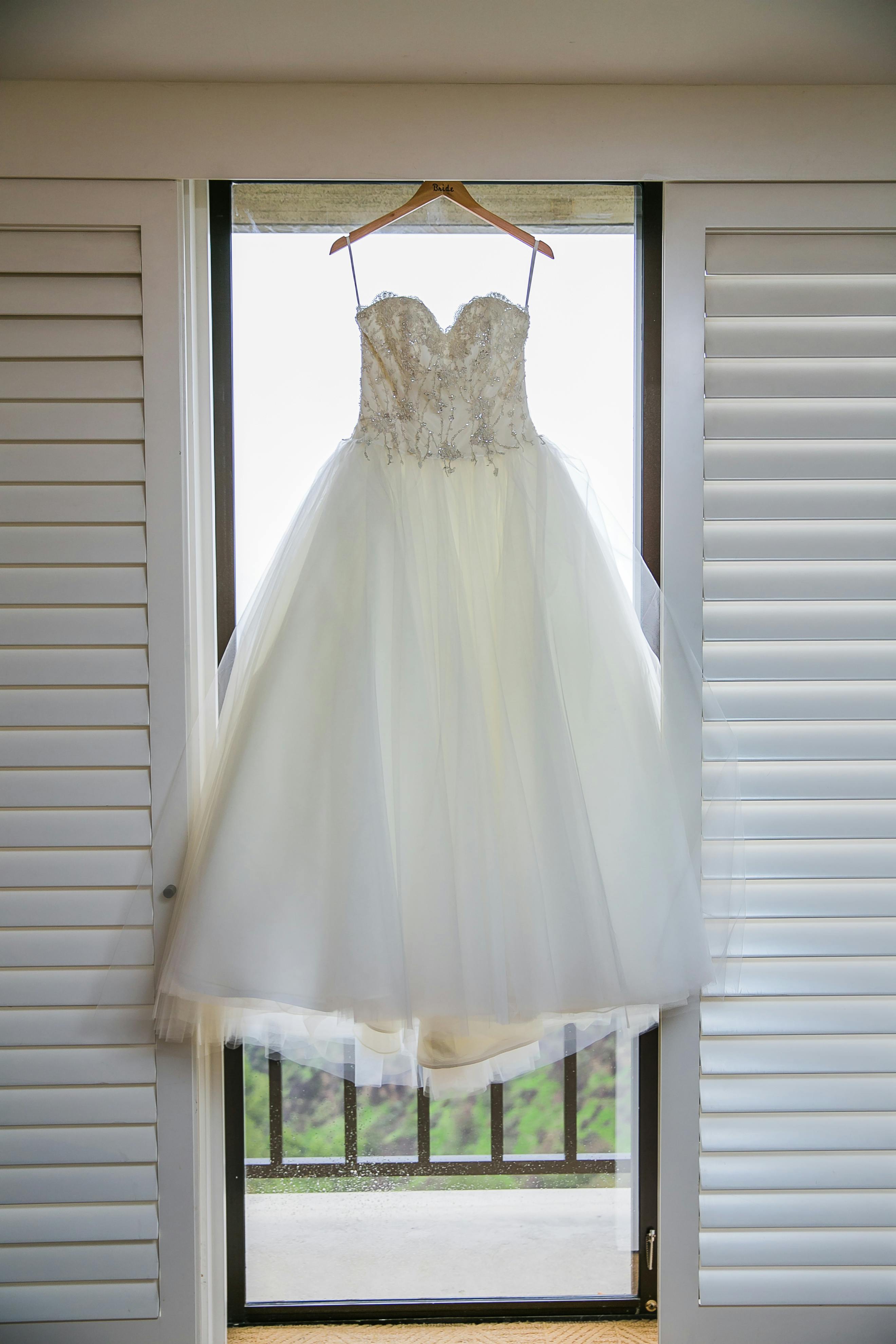 A stunning white bridal gown hangs gracefully in front of a window.
