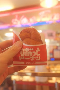 Hand holding a Japanese donut in a vibrant neon-lit restaurant setting.