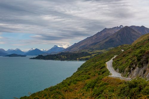 Free A stunning view of a winding road beside a turquoise lake and majestic mountains under a cloudy sky. Stock Photo