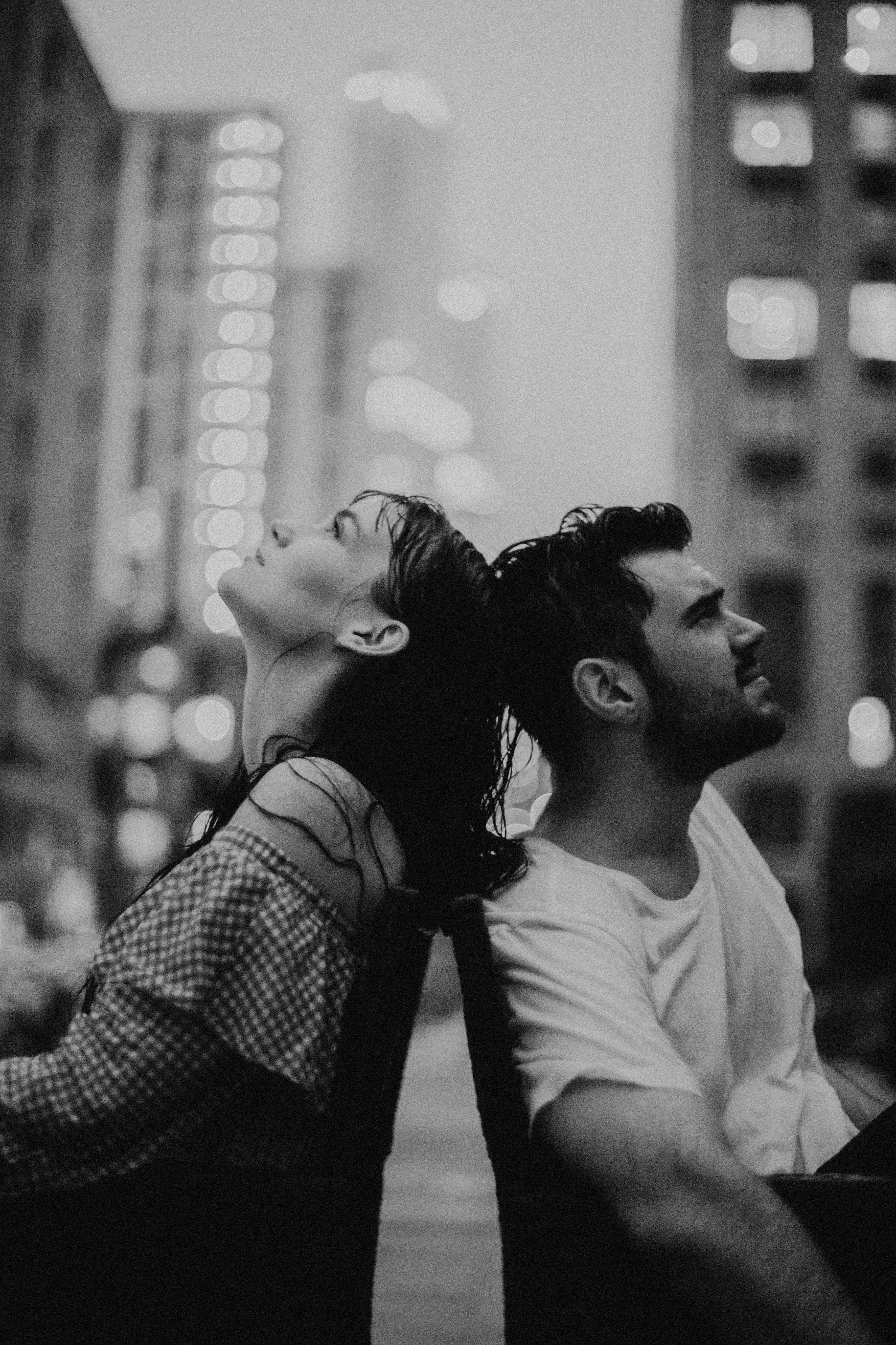 Artistic black and white portrait of a couple sitting back to back in a city setting at night.
