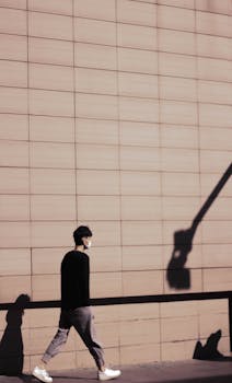 A man wearing a mask walks by a modern building, casting shadows on the wall.