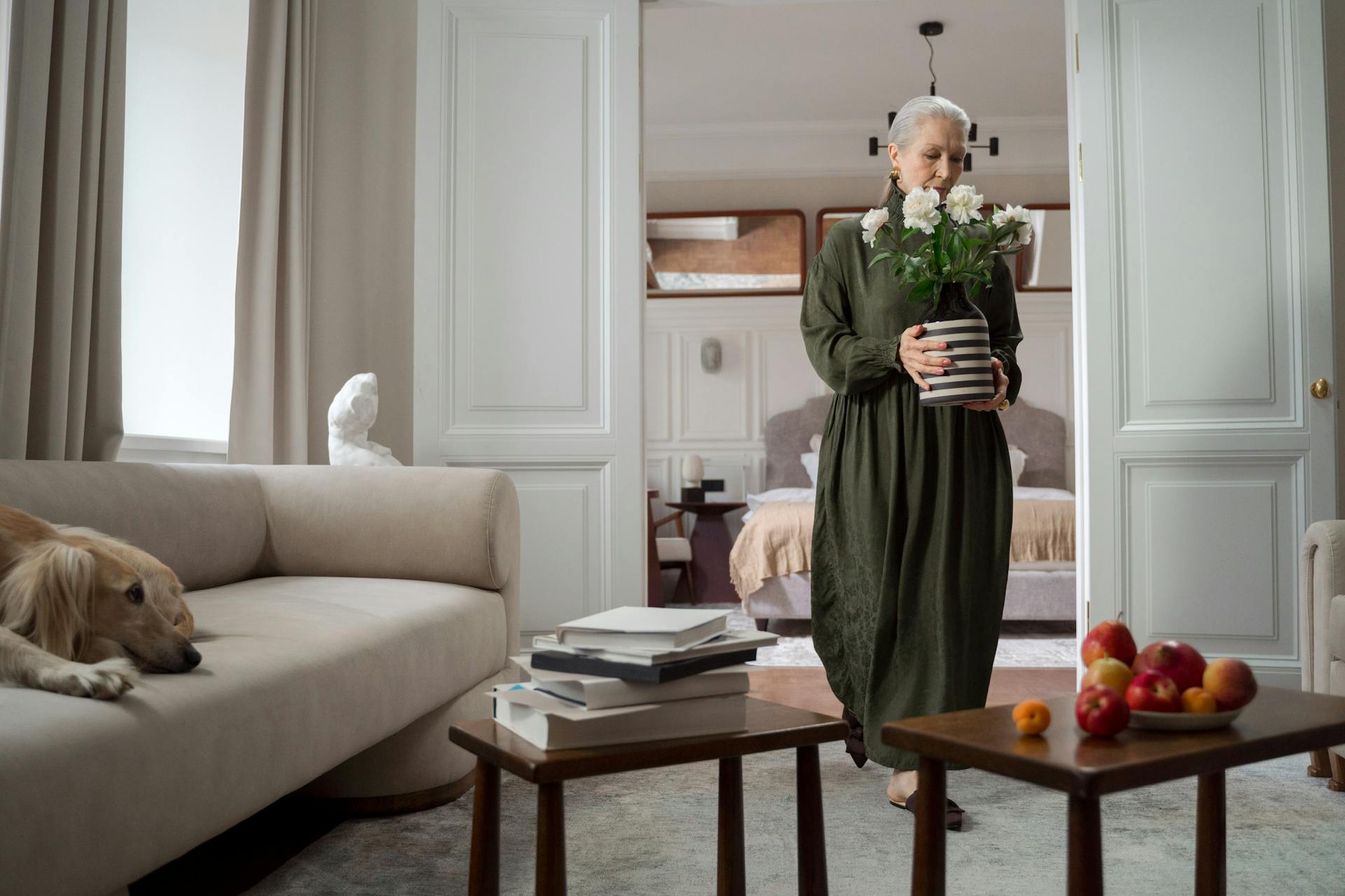 Elderly Woman Carrying Vase with Flowers with Greyhound Dog Resting on Sofa